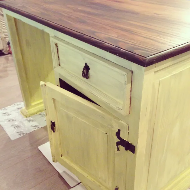 Close-up of a pale yellow distressed wood cabinet with dark wood top and open door showing dark interior; warm rustic mood.